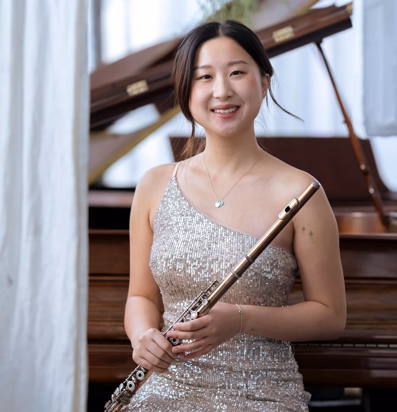 A smiling woman in a sparkly dress holds a flute, standing in front of an open piano with soft lighting and curtains in the background.